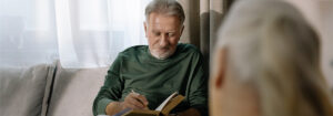 An elderly man in green sweater sitting on the couch while writing in a Book