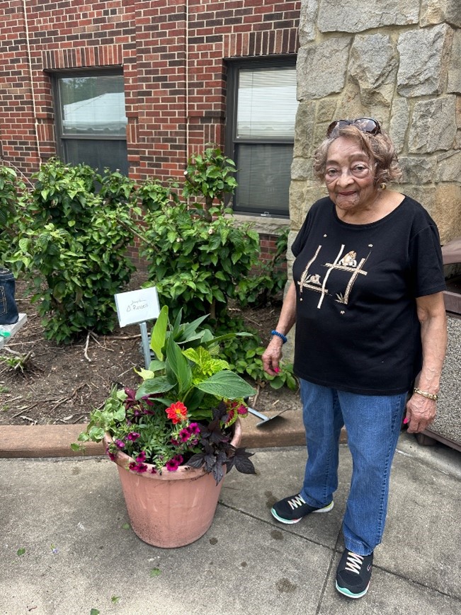 An elderly woman stands beside a potted plant with a small sign, possibly involved in therapeutic gardening for seniors.