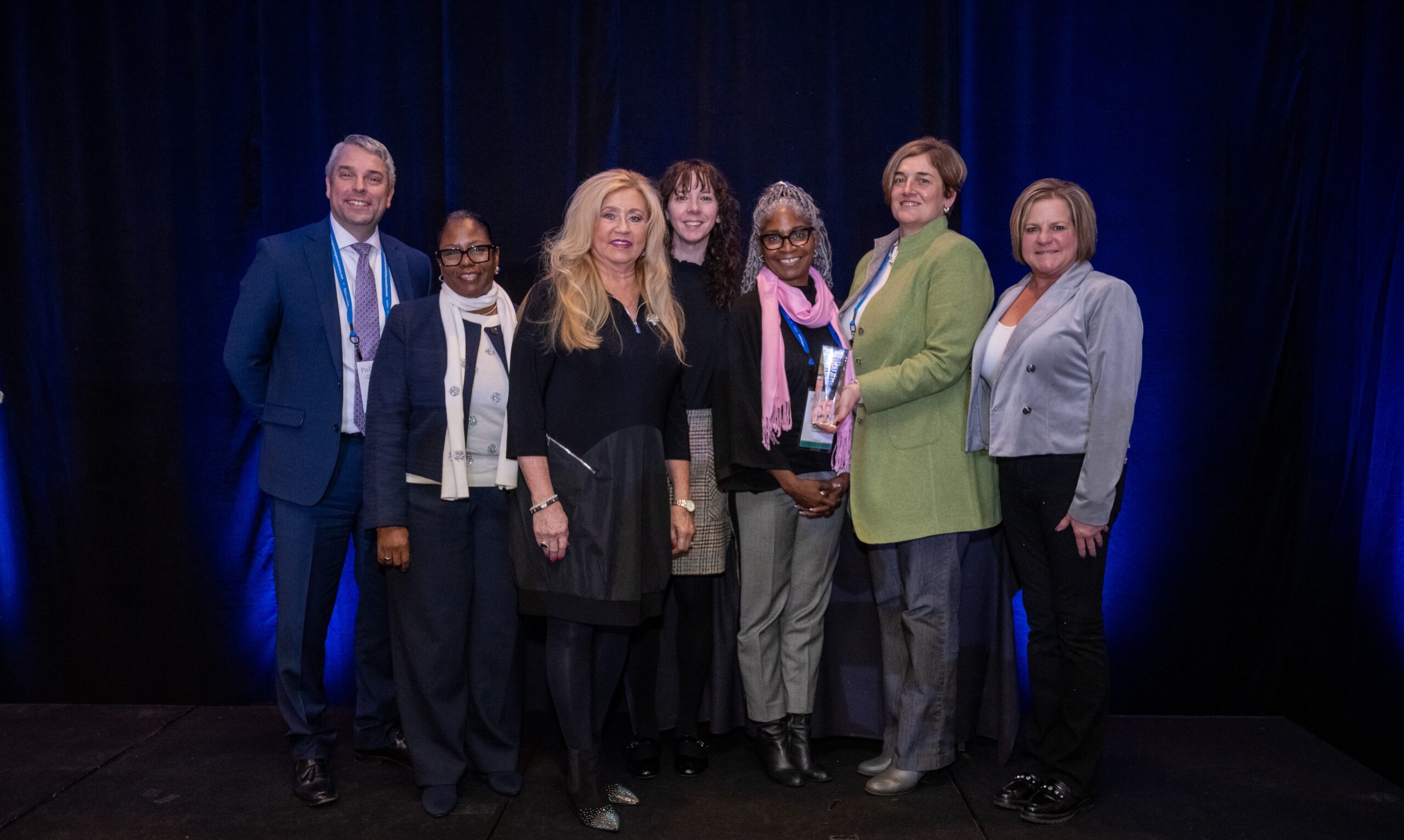 A group of people stands together on a stage with a dark background, posing for a group photo.