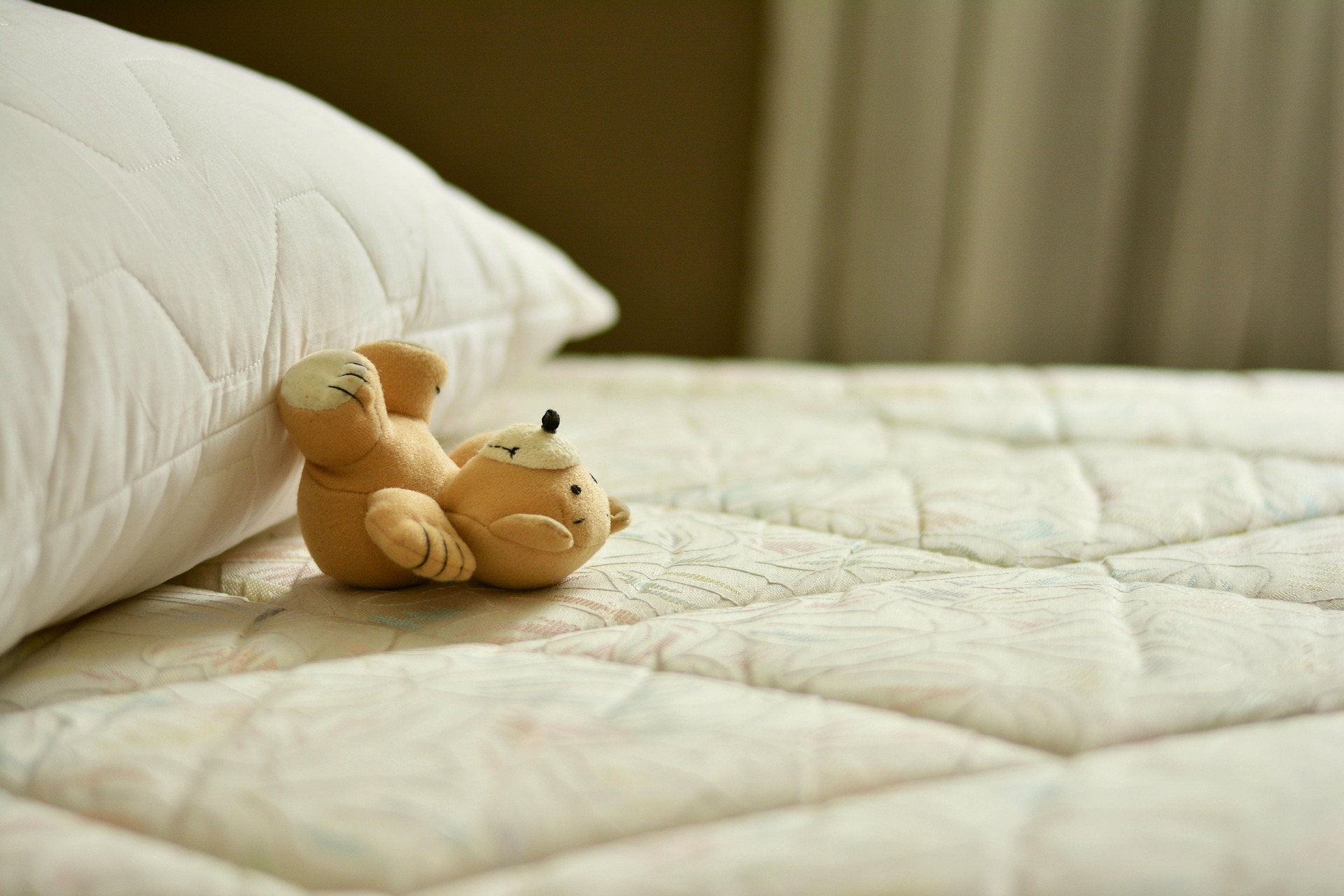 A plush bear lies on a patterned bed near a white pillow, suggesting a cozy and restful setting.